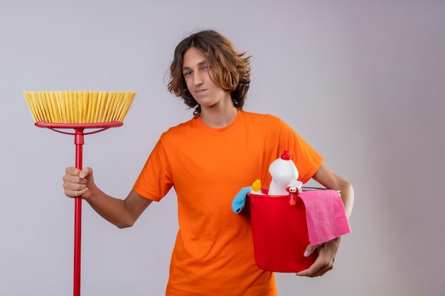 Hombre joven en camiseta naranja sosteniendo un balde con herramientas de limpieza y un trapeador mirando a la cámara con una sonrisa de confianza en la cara de pie sobre fondo blanco.