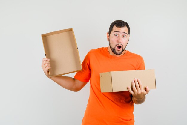 Hombre joven en camiseta naranja sosteniendo una caja de cartón abierta y mirando sorprendido, vista frontal.