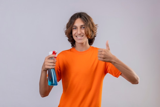 Hombre joven en camiseta naranja con spray de limpieza sonriendo alegremente mirando a la cámara mostrando los pulgares para arriba sobre fondo blanco.
