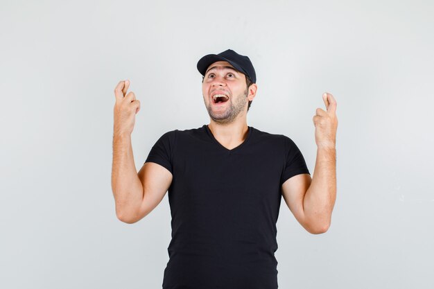 Hombre joven en camiseta negra, gorra sosteniendo los dedos cruzados y mirando alegre