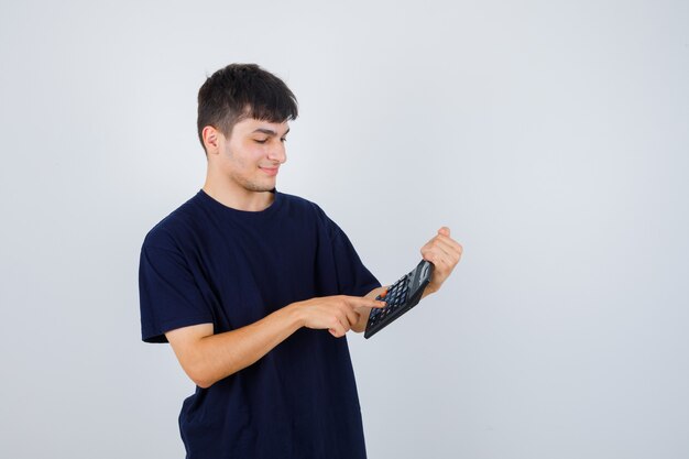 Hombre joven en camiseta negra haciendo cálculos en la calculadora y mirando ocupado, vista frontal.