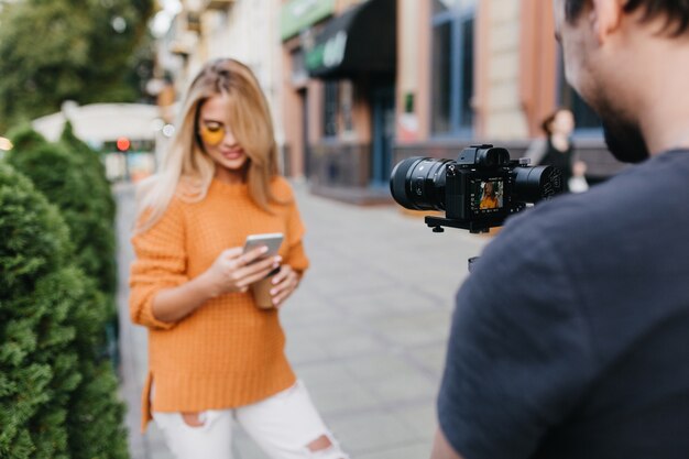 Hombre joven en camiseta negra haciendo foto de mujer rubia alegre en suéter naranja