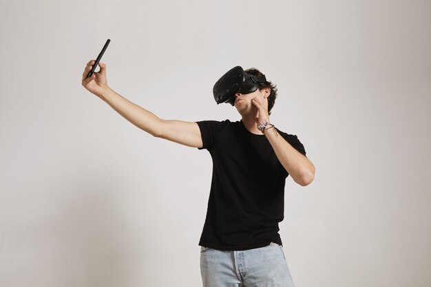 Un hombre joven con camiseta negra y jeans con un auricular VR hace una cara de pato mientras toma una selfie con su teléfono inteligente, aislado en blanco