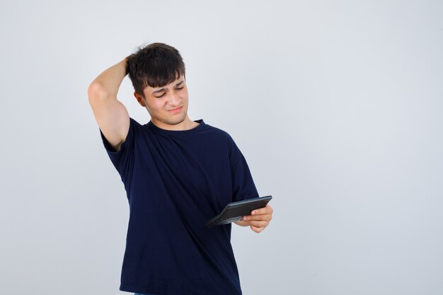 Hombre joven en camiseta negra mirando la calculadora, manteniendo la mano detrás de la cabeza y mirando preocupado, vista frontal.