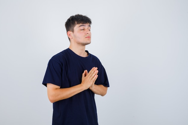 Foto gratuita hombre joven en camiseta negra mostrando gesto de namaste y mirando esperanzado, vista frontal.
