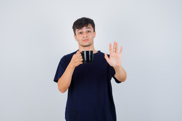 Hombre joven en camiseta negra sosteniendo una taza de té, mostrando gesto de parada y mirando asustado, vista frontal.