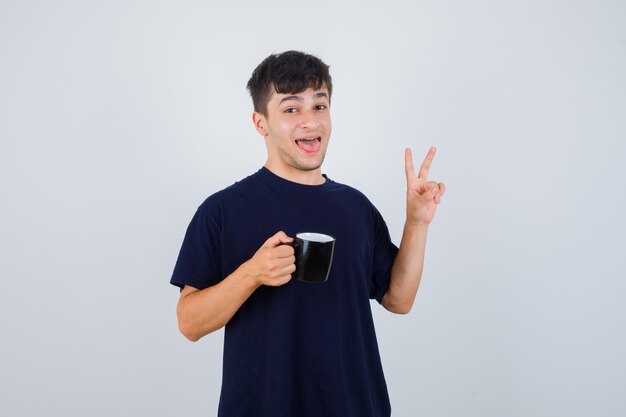 Hombre joven en camiseta negra sosteniendo una taza de té, mostrando el signo V y mirando feliz, vista frontal.