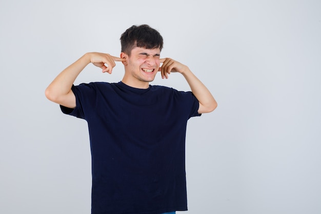 Hombre joven en camiseta negra tapando los oídos con los dedos y mirando molesto, vista frontal.