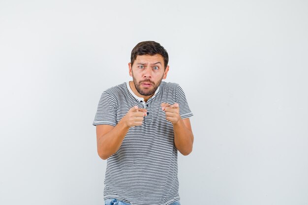 Hombre joven en camiseta a rayas apuntando hacia adelante y mirando perplejo, vista frontal.