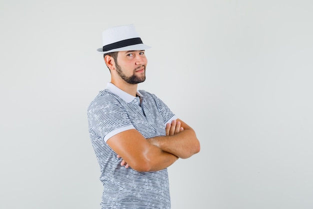 Hombre joven en camiseta a rayas, sombrero mientras está de pie con los brazos cruzados y mirando confiado, vista frontal.