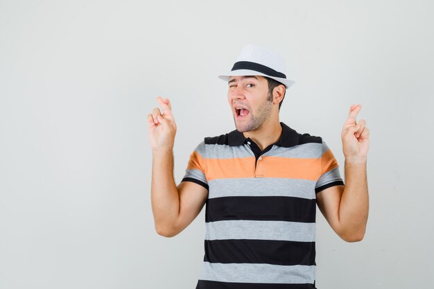 Hombre joven en camiseta a rayas, sombrero de pie con los dedos cruzados y mirando loco espacio para texto