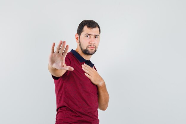 Hombre joven en camiseta roja mostrando gesto de parada mientras sostiene la mano en el pecho y mira seria, vista frontal.