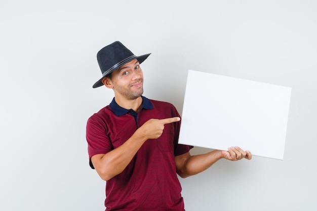 Hombre joven en camiseta, sombrero apuntando al lienzo en blanco y mirando alegre, vista frontal.