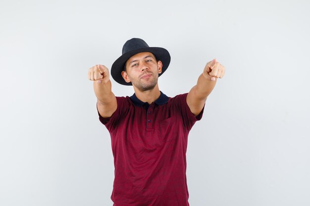 Hombre joven en camiseta, sombrero apuntando a la cámara y mirando confiado, vista frontal.