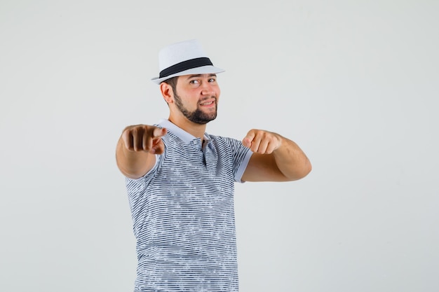 Hombre joven en camiseta, sombrero apuntando a la cámara y mirando seguro, vista frontal.