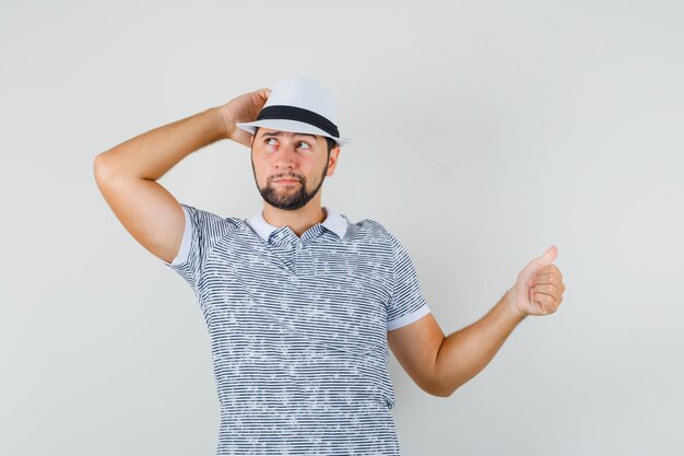 Hombre joven en camiseta, sombrero apuntando con el pulgar hacia un lado y mirando pensativo, vista frontal.