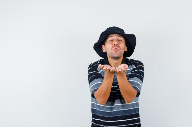 Hombre joven en camiseta, sombrero enviando beso que sopla, vista frontal.