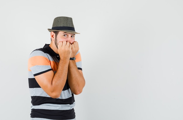 Hombre joven en camiseta, sombrero mordiendo los puños emocionalmente y mirando asustado, vista frontal.