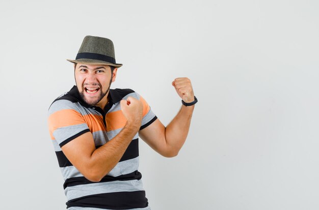 Hombre joven en camiseta, sombrero mostrando gesto de ganador y con suerte, vista frontal.