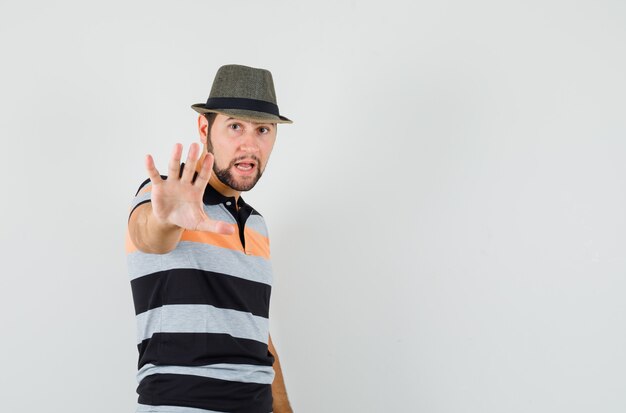 Hombre joven en camiseta, sombrero mostrando gesto de parada y mirando serio, vista frontal.