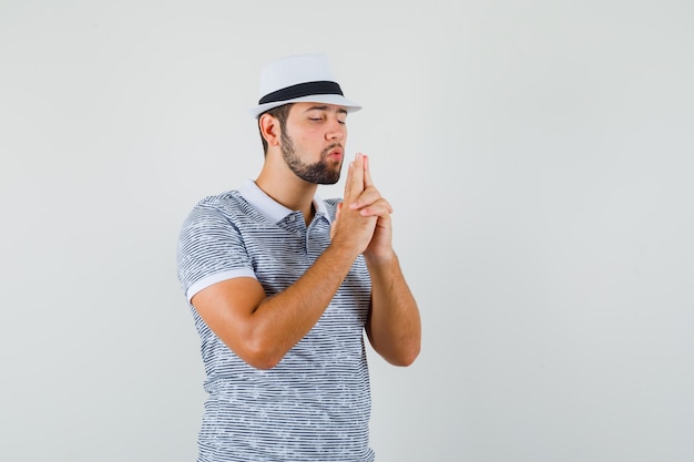 Hombre joven en camiseta, sombrero mostrando gesto de pistola y mirando intrépido, vista frontal.