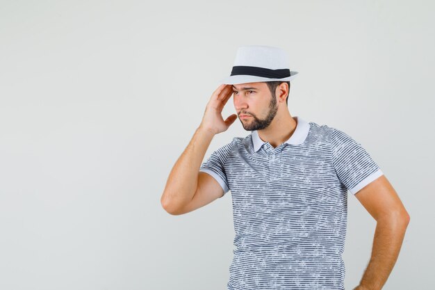 Hombre joven en camiseta, sombrero tirando de la piel de sus sienes y mirando pensativo, vista frontal.