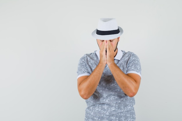 Hombre joven en camiseta, sombrero tomados de la mano en gesto de oración y mirando en silencio, vista frontal.