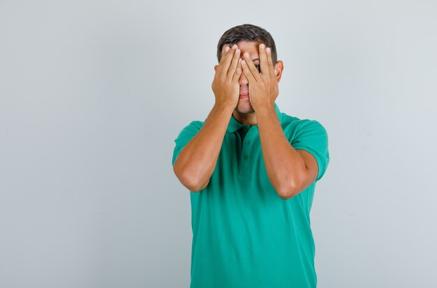 Hombre joven en camiseta verde cubriendo sus ojos y mirando lo siento, vista frontal.