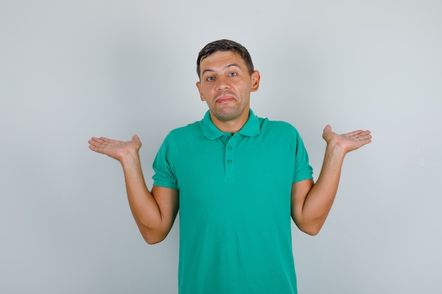 Hombre joven en camiseta verde que muestra un gesto de impotencia y parece confundido, vista frontal.