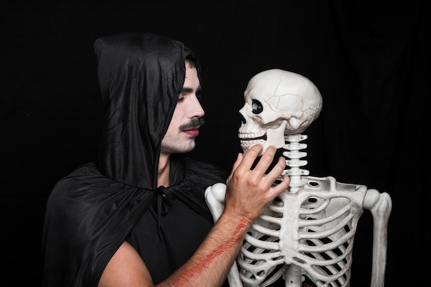 Hombre joven en capa negra con capucha mirando el esqueleto