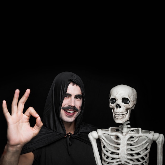 Hombre joven en la capa negra que se coloca con el esqueleto y que muestra gesto aceptable