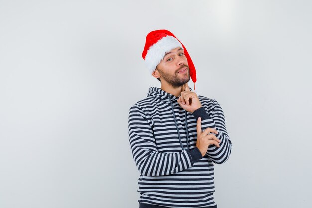 Hombre joven con capucha, sombrero de Santa de pie en pose de pensamiento y mirando inteligente, vista frontal.