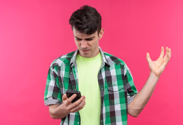 Hombre joven caucásico enojado con camisa verde mirando el teléfono en la mano y la mano levantada en la pared rosa aislada