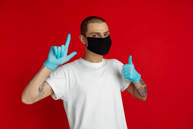 Hombre joven caucásico con mascarilla protectora y guantes médicos en la pared roja muestra el pulgar hacia arriba, apuntando. Concepto de emociones humanas, ventas, bloqueo por coronavirus, publicidad, cuarentena, aislamiento.