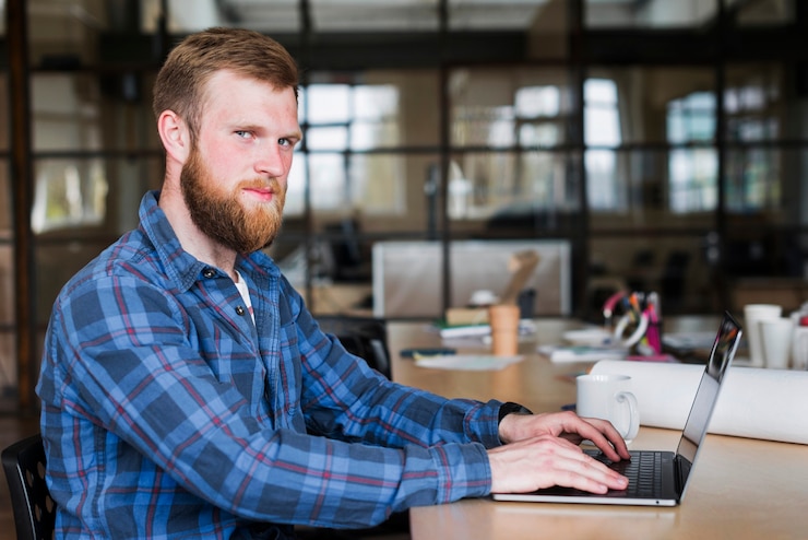 Foto gratuito hombre joven caucásico que se sienta delante de la computadora portátil y que mira la cámara