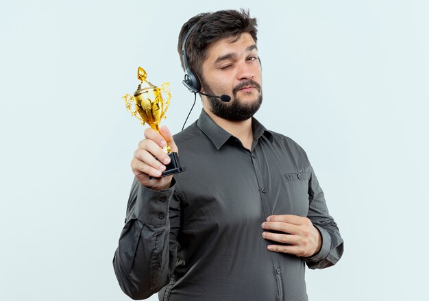 Hombre joven del centro de llamadas parpadeó con auriculares sosteniendo la copa ganador aislado sobre fondo blanco.