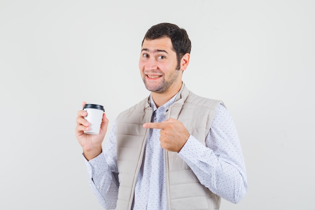 Hombre joven con chaqueta beige apuntando a la taza de café para llevar con el dedo índice y mirando optimista, vista frontal.