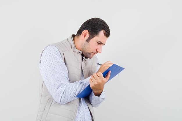 Hombre joven en chaqueta beige escribiendo algo en el cuaderno con lápiz y mirando enfocado, vista frontal.