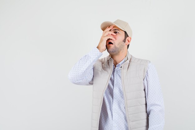 Hombre joven con chaqueta beige y gorra cubriendo parte de la cara con la mano y mirando sorprendido, vista frontal.