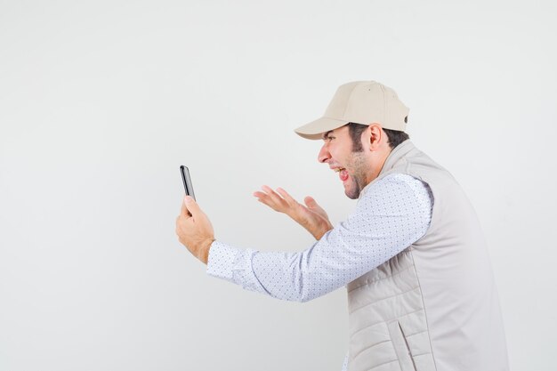 Hombre joven con chaqueta beige y gorra hablando con alguien a través de una videollamada y sacando la lengua y mirando divertido, vista frontal.