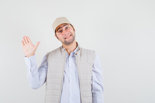 Hombre joven con chaqueta beige y gorra levantando la mano saludando a alguien y mirando feliz, vista frontal.