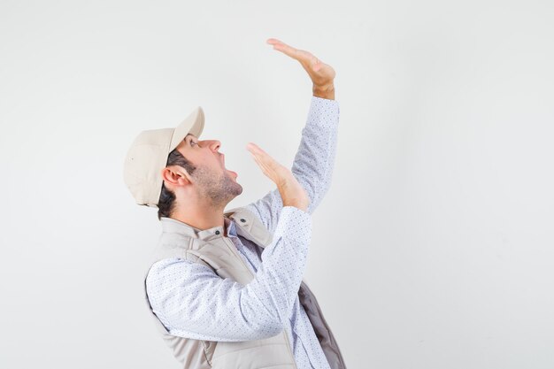 Hombre joven con chaqueta beige y gorra levantando las manos como sosteniendo algo y mirando sorprendido, vista frontal.