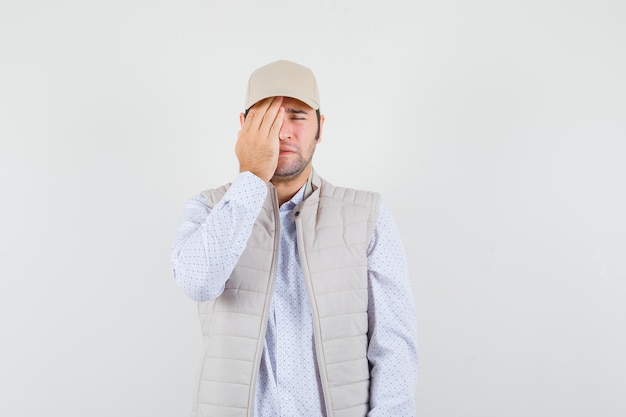 Hombre joven con chaqueta beige y gorra que cubre los ojos con la mano y parece cansado, vista frontal.