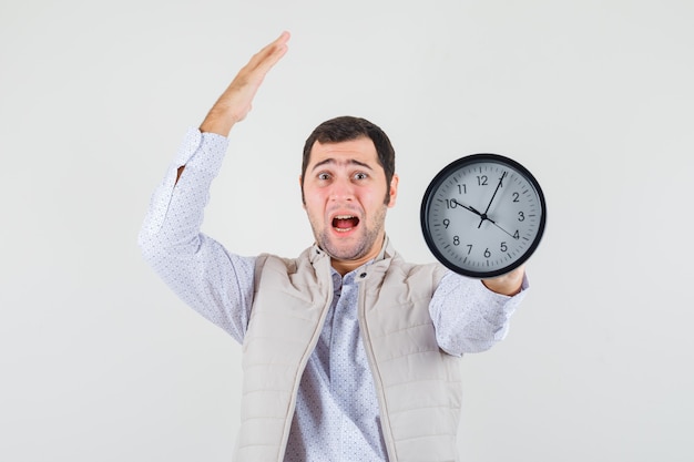 Hombre joven con chaqueta beige y gorra sosteniendo el reloj en uno y estirando la mano por encima de la cabeza y mirando sorprendido, vista frontal.