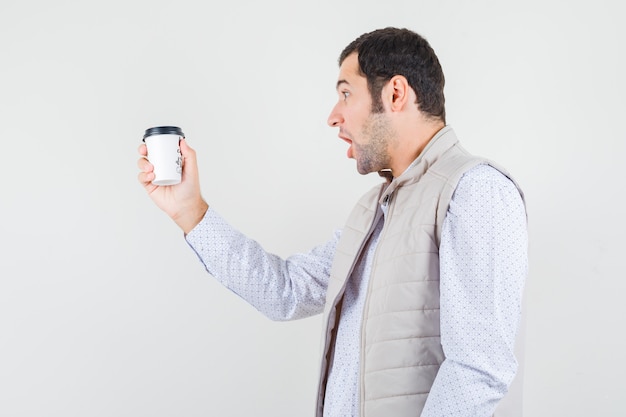 Foto gratuita hombre joven con chaqueta beige sosteniendo una taza de café para llevar y mirándolo y mirando sorprendido, vista frontal.