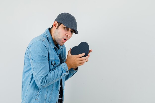 Hombre joven con chaqueta, gorra agitando caja de regalo y mirando emocionado espacio para texto