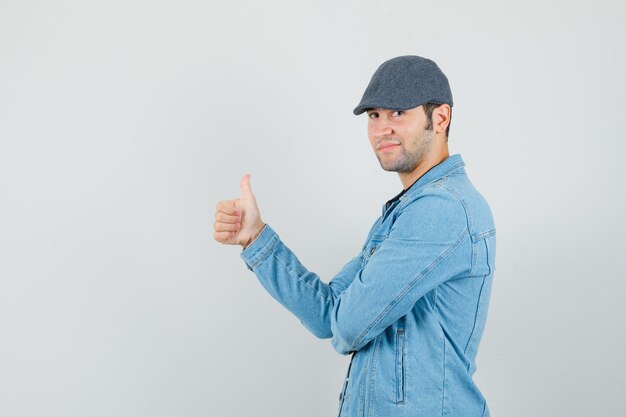 Hombre joven con chaqueta, gorra mostrando el pulgar hacia arriba y mirando confiado.