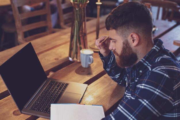 Hombre joven charlando a través de net-book durante el descanso del trabajo en la cafetería, hombre sentado frente a una computadora portátil abierta con pantalla de espacio de copia en blanco para su mensaje de texto o contenido publicitario