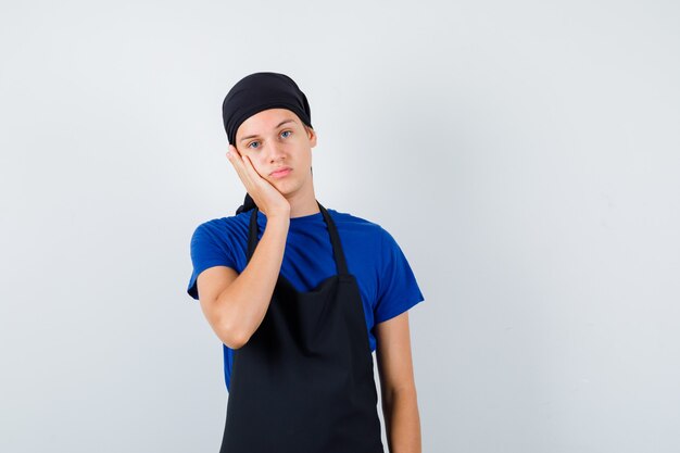 Hombre joven cocinero en camiseta, delantal apoyado en la mejilla en la mano y mirando pensativo, vista frontal.