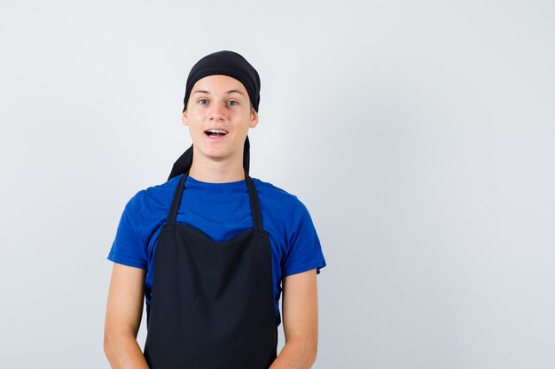 Hombre joven cocinero en camiseta, delantal posando mientras está de pie y mirando feliz, vista frontal.